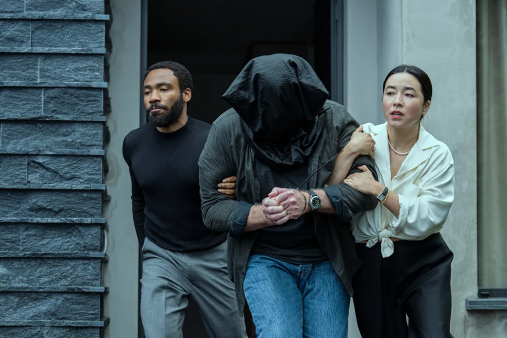Maya Erskine and Donald Glover as Jane and John Smith in Mr. and Mrs. Smith. With Ron Perlman. Image courtesy of Prime Video.